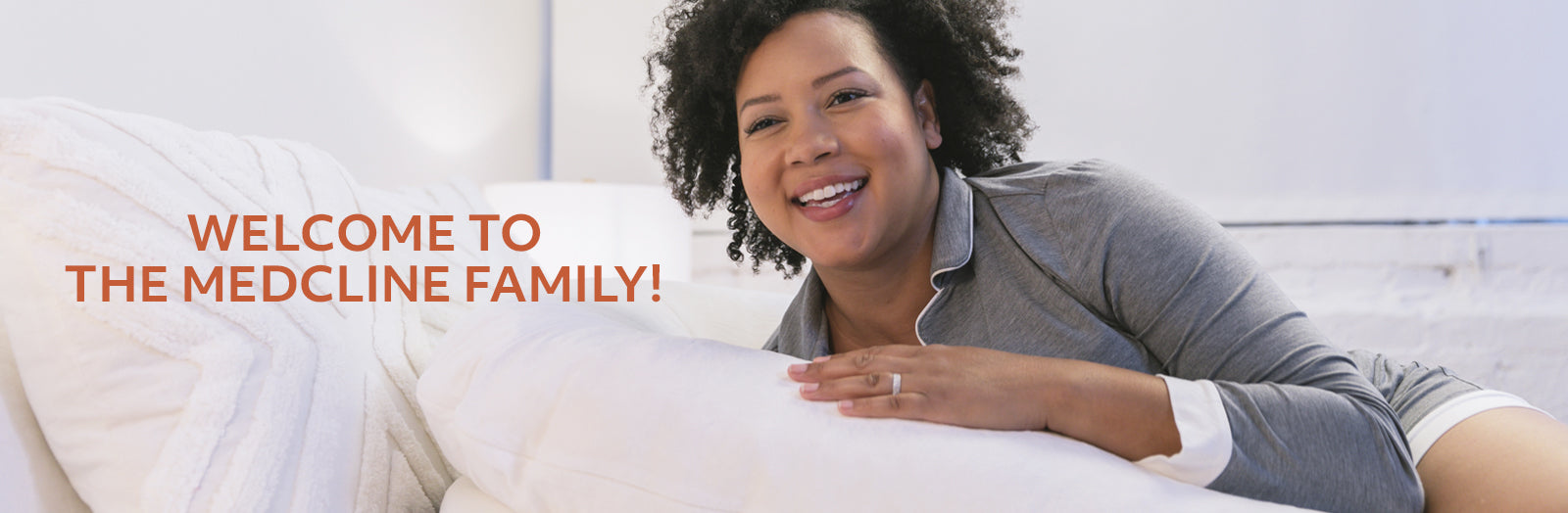 A smiling woman in gray pajamas sitting on a bed, leaning on a therapeutic body pillow.