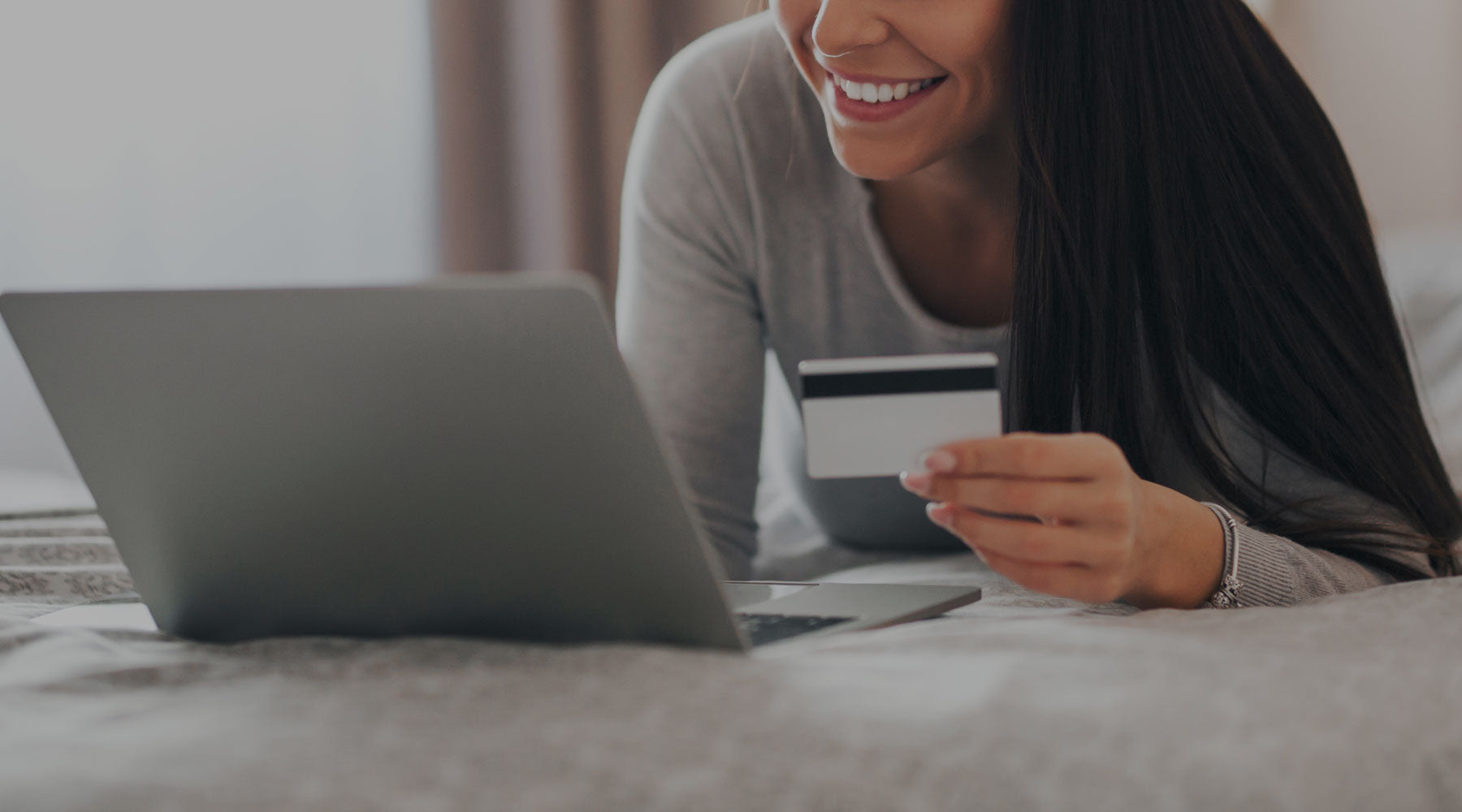 Smiling woman holding a credit card while shopping online on a laptop from the comfort of her bed.